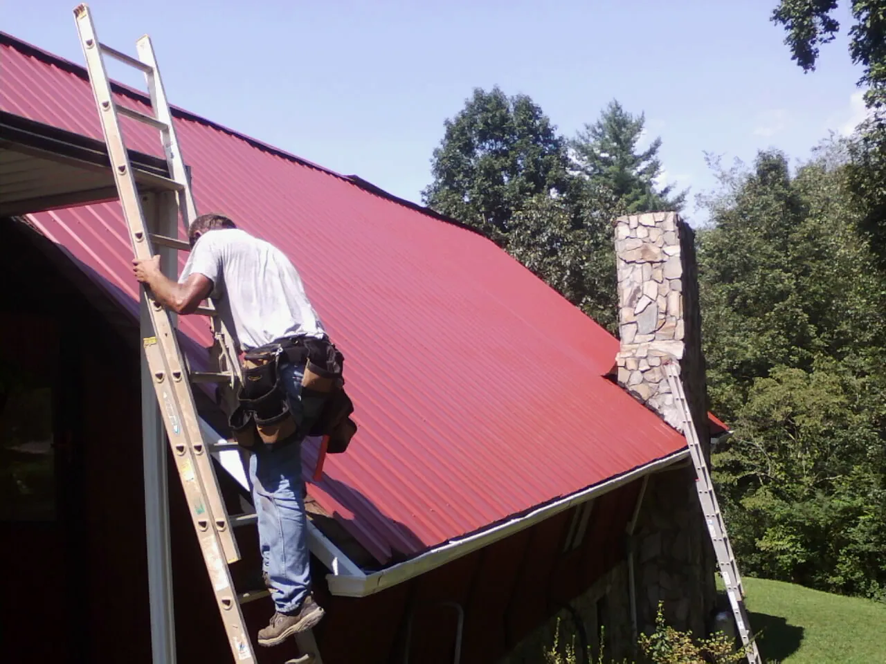 Roofer on a ladder beside a red metal roof and stone chimney — Soffit & Fascia Repair by Roofing Brothers in Longmeadow
